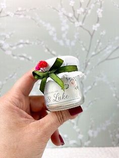 a hand holding a small glass jar with a flower on it's side and a green ribbon around the top