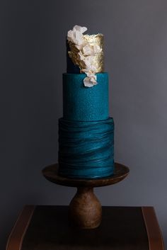 a three tiered blue cake with gold and white flowers on top, sitting on a wooden stand