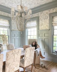 a woman sitting at a dining room table in front of a chandelier and windows