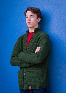 a young man leaning against a blue wall wearing a green cardigan sweater and jeans