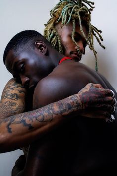 two men with dreadlocks hugging each other in front of a white wall and one has tattoos on his arm