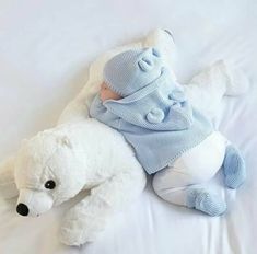 a white teddy bear laying on top of a bed next to a baby wearing a blue sweater and hat