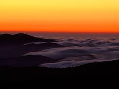 the sun is setting over some mountains covered in fog and clouds with an airplane flying overhead