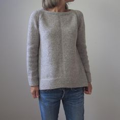 a woman standing in front of a white wall wearing a gray sweater and blue jeans