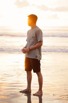 a man standing on the beach at sunset with his hand in his pocket looking out to sea