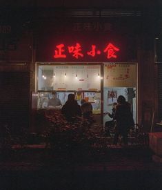 two people are standing in front of a restaurant with red neon lights on the windows