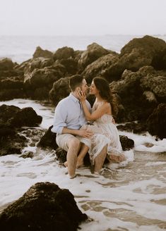 a man and woman are sitting on rocks in the water at the beach while holding each other's hands