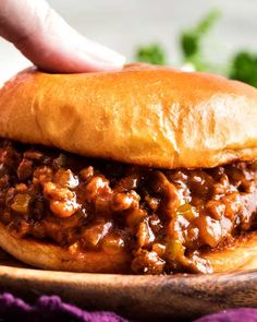 a hand is picking up a sloppy joe sandwich from a wooden plate on a purple cloth