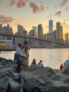 people are sitting on the rocks near the water and in front of a city skyline