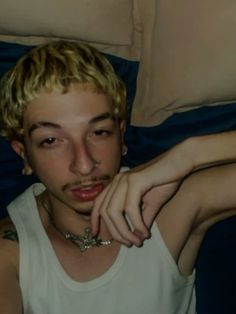 a young man with blonde hair wearing a white tank top sitting on a bed next to pillows