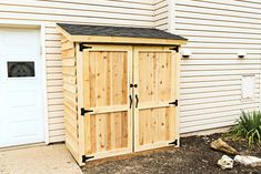 a wooden storage shed sitting in front of a white building with two doors on each side