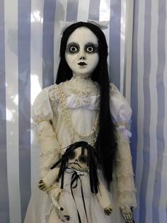 a creepy doll with long black hair and white dress is standing in front of striped wallpaper