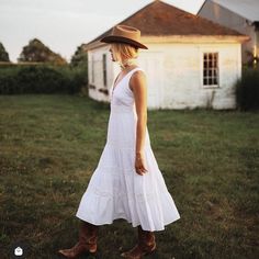 a woman in a white dress and brown hat standing on the grass near an old house