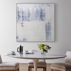 a painting hanging above a table with two chairs and a vase filled with flowers on it