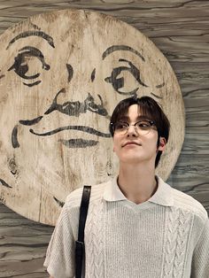 a young man wearing glasses standing in front of a wooden wall with a face drawn on it