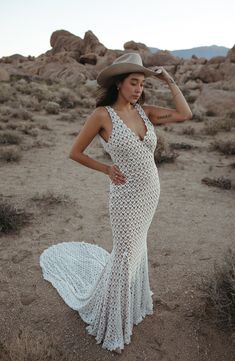 a woman standing in the desert wearing a white dress and cowboy hat with her hands on her head