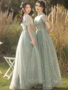 two young women dressed in dresses standing next to each other on the grass with tall grasses behind them