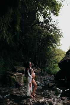 a pregnant woman standing in the middle of a stream with trees around her and rocks on either side