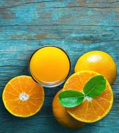 three oranges and one glass of juice on a blue wooden table with green leaves