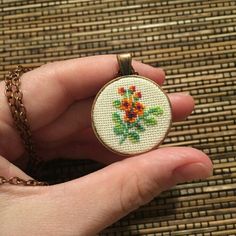 a hand is holding a small cross stitched necklace with flowers on it and a gold chain