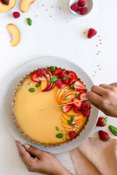 a person cutting into a cheesecake topped with sliced peaches and strawberries on a white table