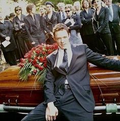 a man in a suit and tie sitting on the hood of a car with other people behind him