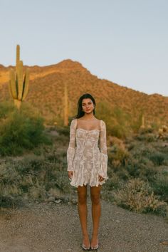 a woman standing in the middle of a desert with a cactus and mountains behind her