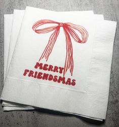 two white napkins with red ribbon and merry friends written on the front one has a bow