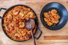 two pans filled with shrimp and rice on top of a wooden table next to a spoon