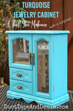 a turquoise jewelry cabinet with glass doors and gold knobs on the bottom, sitting on a table