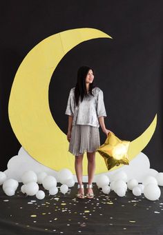 a woman holding a star standing in front of a moon and clouds backdrop with balloons