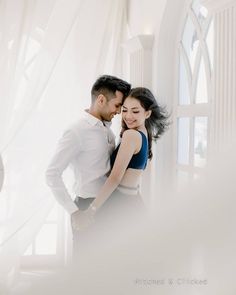 a man and woman standing next to each other in front of a window with sheer curtains