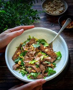 two hands holding a white bowl filled with meat and veggies on top of a wooden table