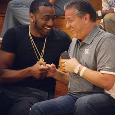 two men sitting next to each other and one is looking at his cell phone while the other man smiles