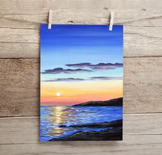 a painting hanging on a clothes line with the sun setting over the ocean in the background