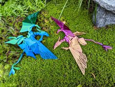 three different colored dragon kites laying on the ground next to some green grass and rocks