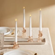 four candles are lit on a table with white plates and gold cutleryware in the foreground