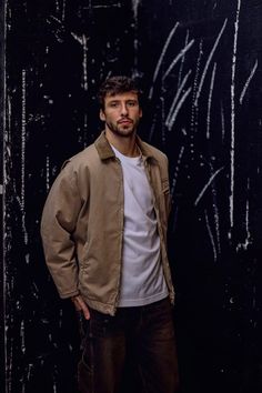 a man standing in front of a black wall wearing a jacket and white t - shirt