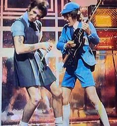 two men in blue uniforms are playing guitar and singing on the television screen while another man is standing behind them