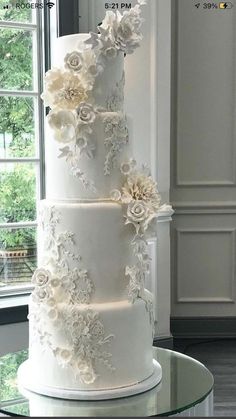 a three tiered white wedding cake with flowers on the top and bottom, sitting on a glass table in front of a window