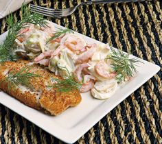 a white plate topped with fish next to potato salad and a fork on top of a woven place mat