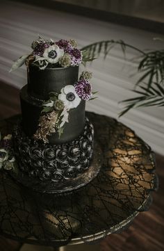 a three tiered black cake with flowers on top and greenery in the background