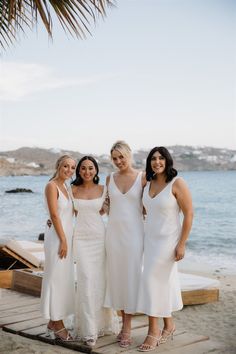 three women in white dresses standing on a dock next to the ocean and palm trees