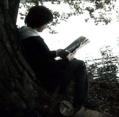 a person sitting on a tree next to a body of water with a book in their hands