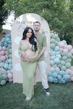 a pregnant couple standing in front of an outdoor backdrop with balloons and flowers on it