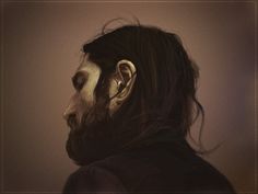 a man with long hair and beard standing in front of a white wall wearing an earring