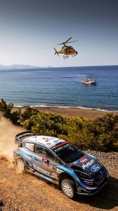 a helicopter flying over a car driving down a dirt road next to the ocean with boats in the background