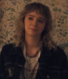 a woman with blonde hair wearing a black jacket and white shirt standing in front of floral wallpaper