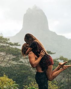 a man holding a woman in his arms while they are standing in the rain with mountains behind them