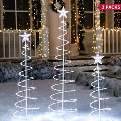 three lighted christmas trees in front of a house with snow on the ground and lights around them